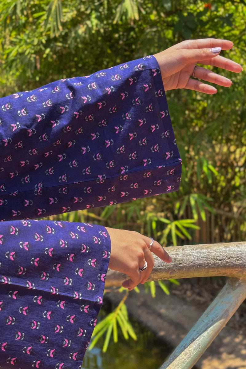 Person wearing a jiva-blue patterned outfit with hands on a railing against a green foliage background 