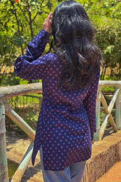 Person wearing a jiva-blue patterned shirt standing on a wooden bridge with greenery in the background 