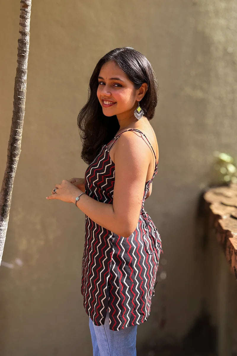 Woman in a zigzag patterned dress standing against a beige wall with a plant to her left.