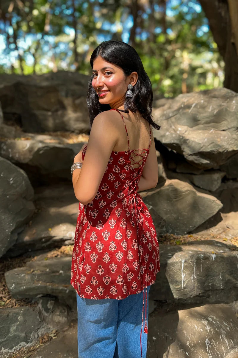 Woman in a butta red patterned top and jeans standing in a natural setting with rocks and trees.