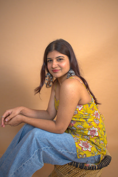 Woman sitting on a woven chair wearing a jal-yellow floral top and blue jeans against a beige background