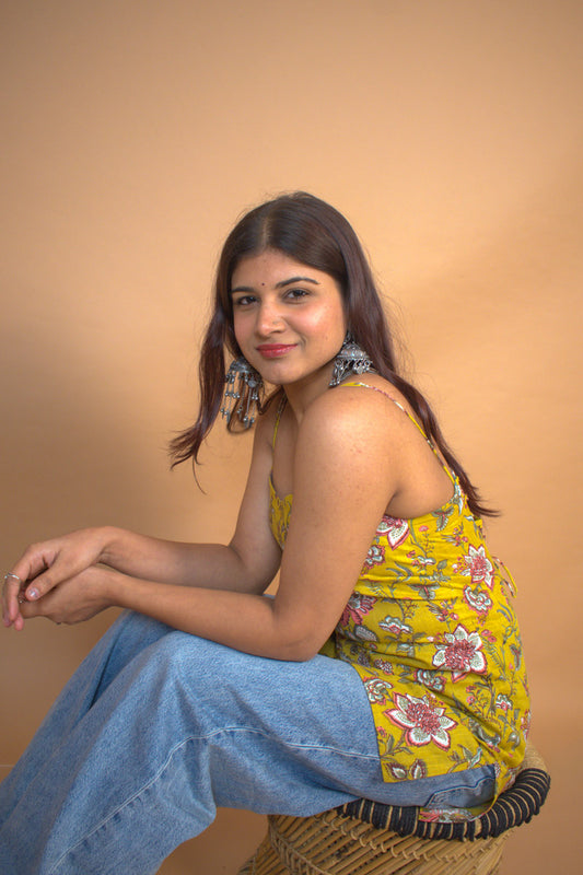 Woman sitting on a woven chair wearing a jal-yellow floral top and blue jeans against a beige background