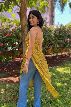 Woman in a jiva-yellow dress and blue jeans standing outdoors with greenery in the background