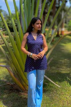 Woman in a jiva-blue top and light blue jeans standing in a garden with large green leaves.
